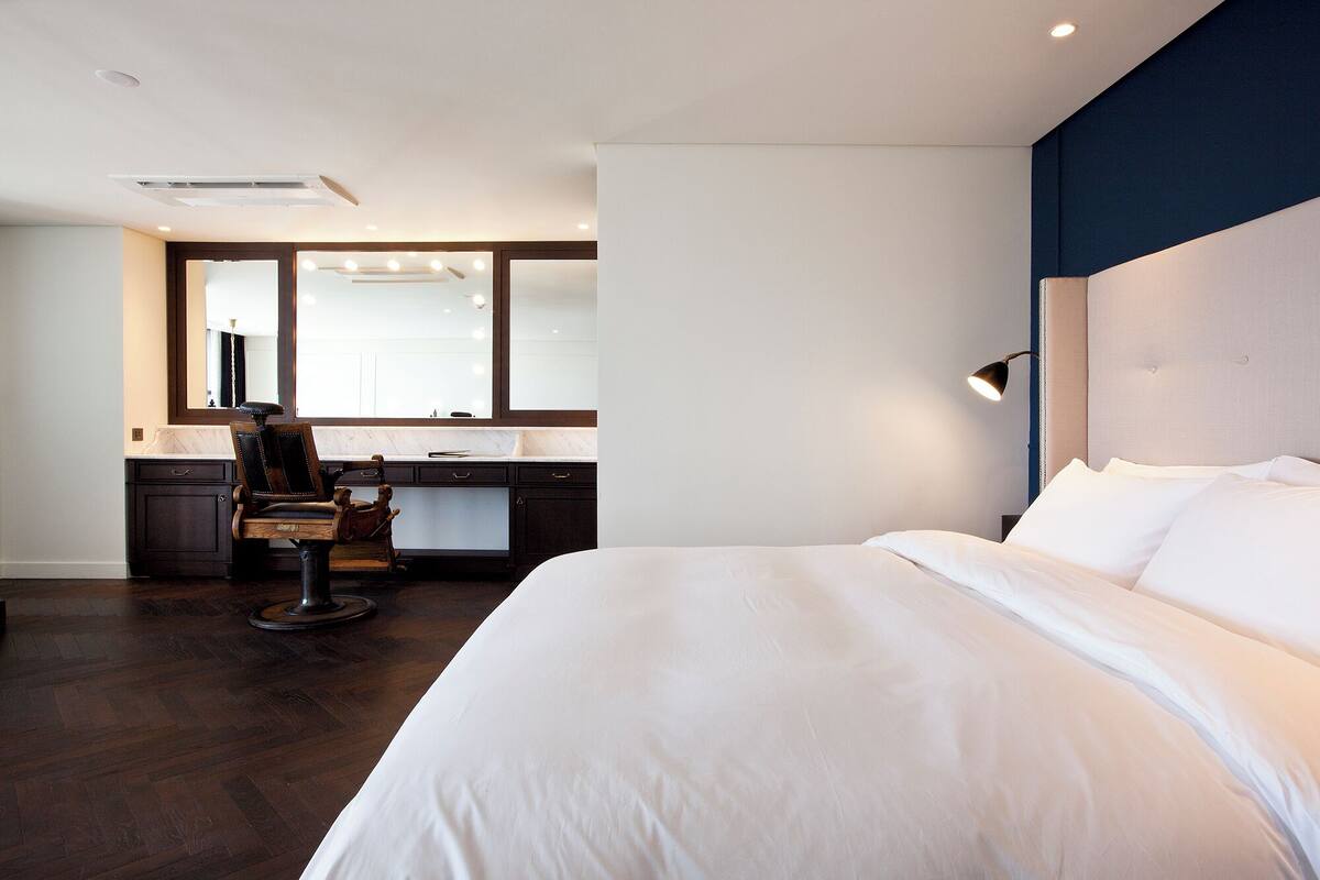A hotel room with a large bed, a desk with a chair, a mirror, and a headboard with a lamp.