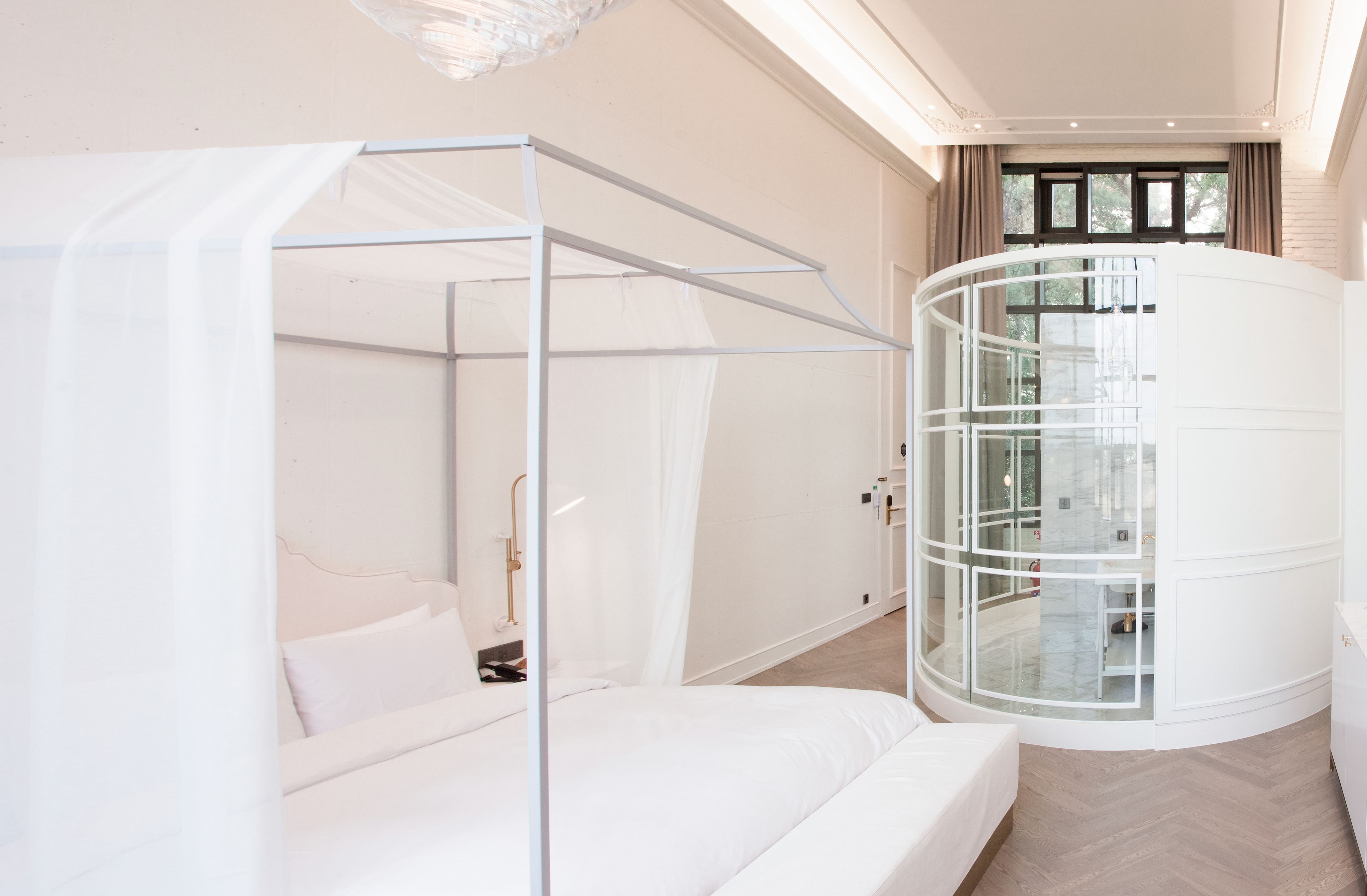 Quarto moderno com cama de dossel, poltrona giratória com estrutura de vidro e vista para o exterior através de uma janela.