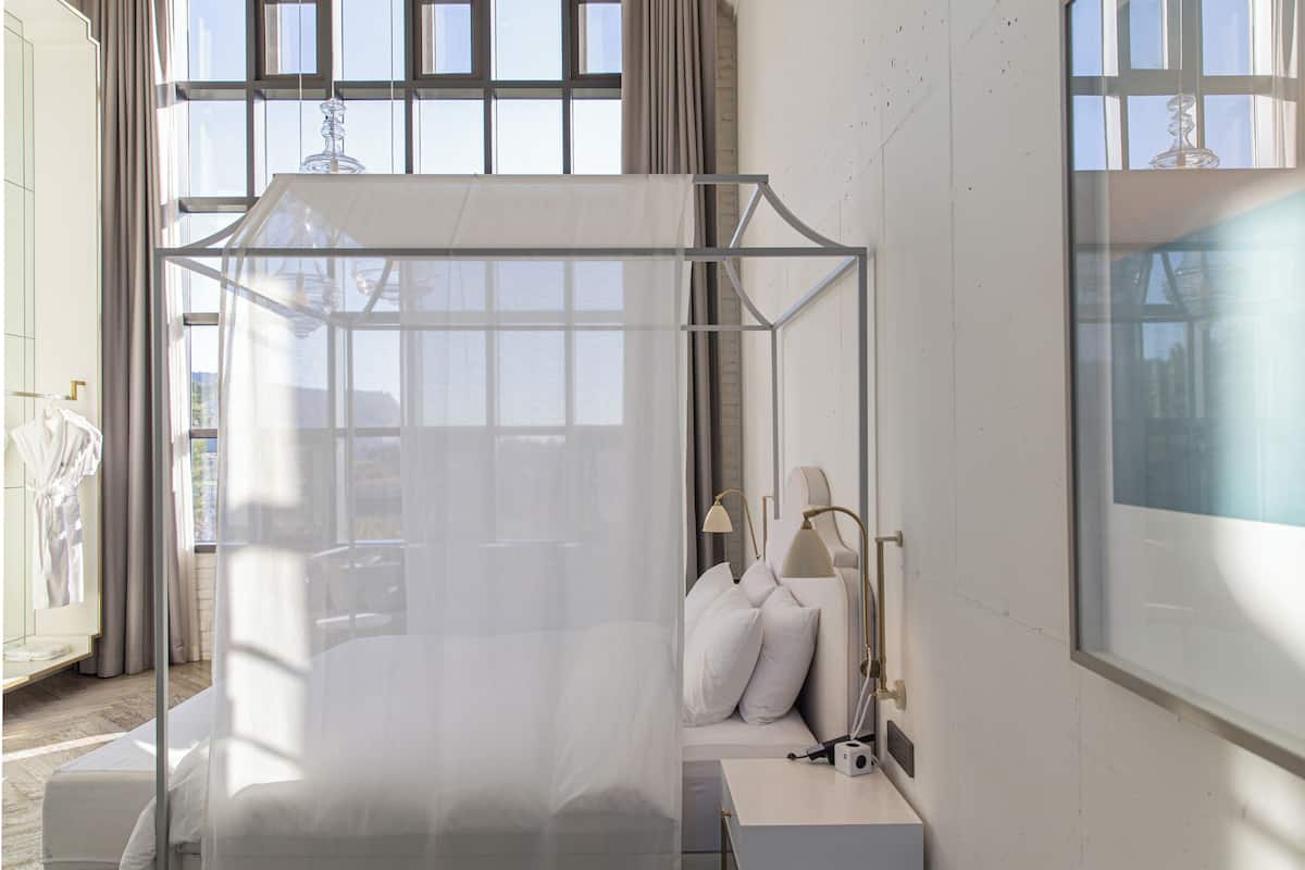 A modern bedroom with a canopy bed, a bedside table, a lamp, and a view of the outside through large windows.