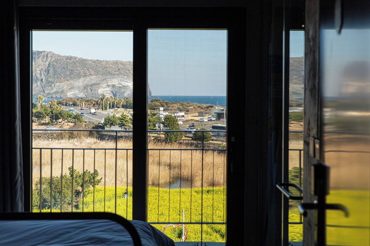 A room with a large window overlooking a coastal landscape, a balcony with a metal railing, and a bed with a white headboard.