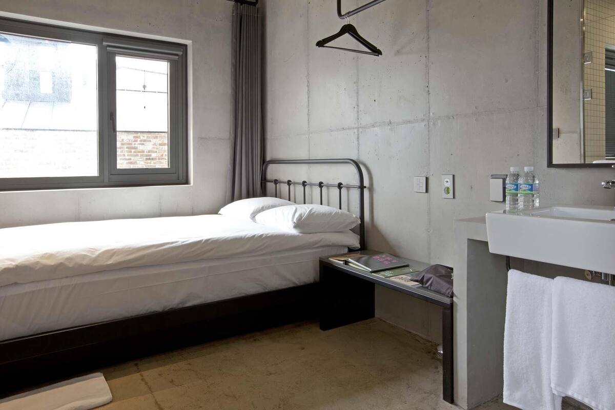 A small, modern hotel room with a single bed, a bedside table with a book, a mirror, and a window with a view of the outside.