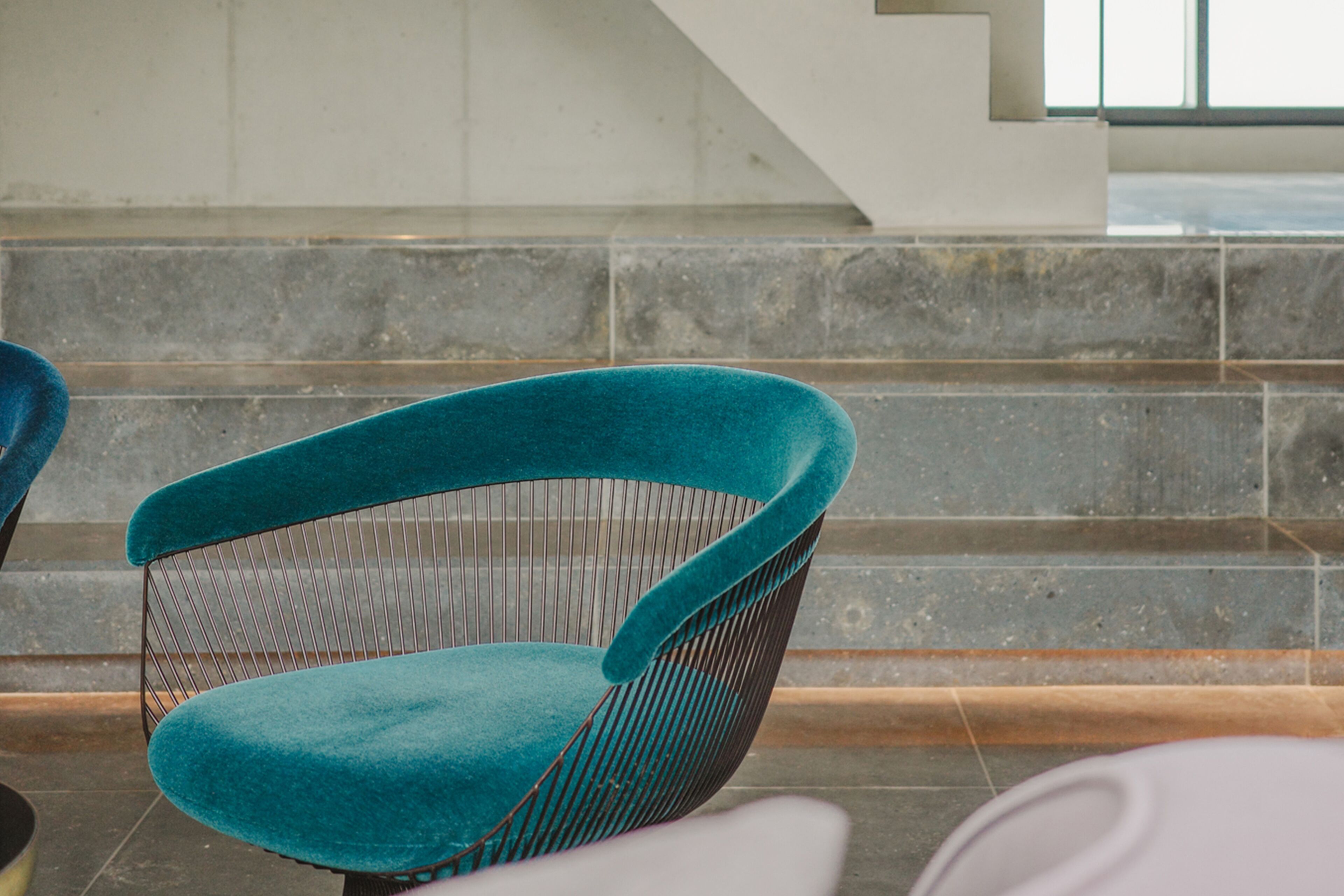 A teal upholstered chair with a wireframe backrest, positioned in front of a staircase with stone steps and a modern interior design.