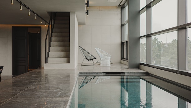 Una piscina interior moderna con vistas despejadas al cielo a través de grandes ventanales, una escalera y dos sillas blancas cerca de la piscina.