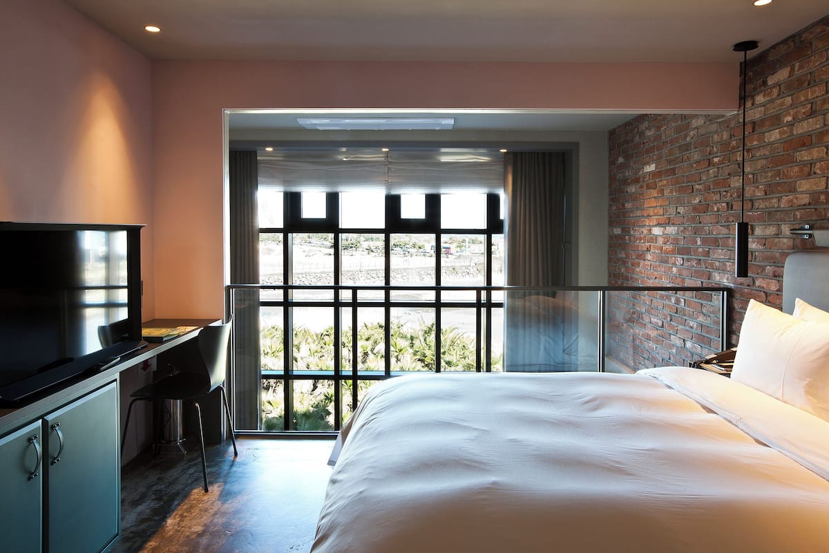 A modern hotel room with a large bed, a desk with a TV, and a view of the outdoors through a large window.