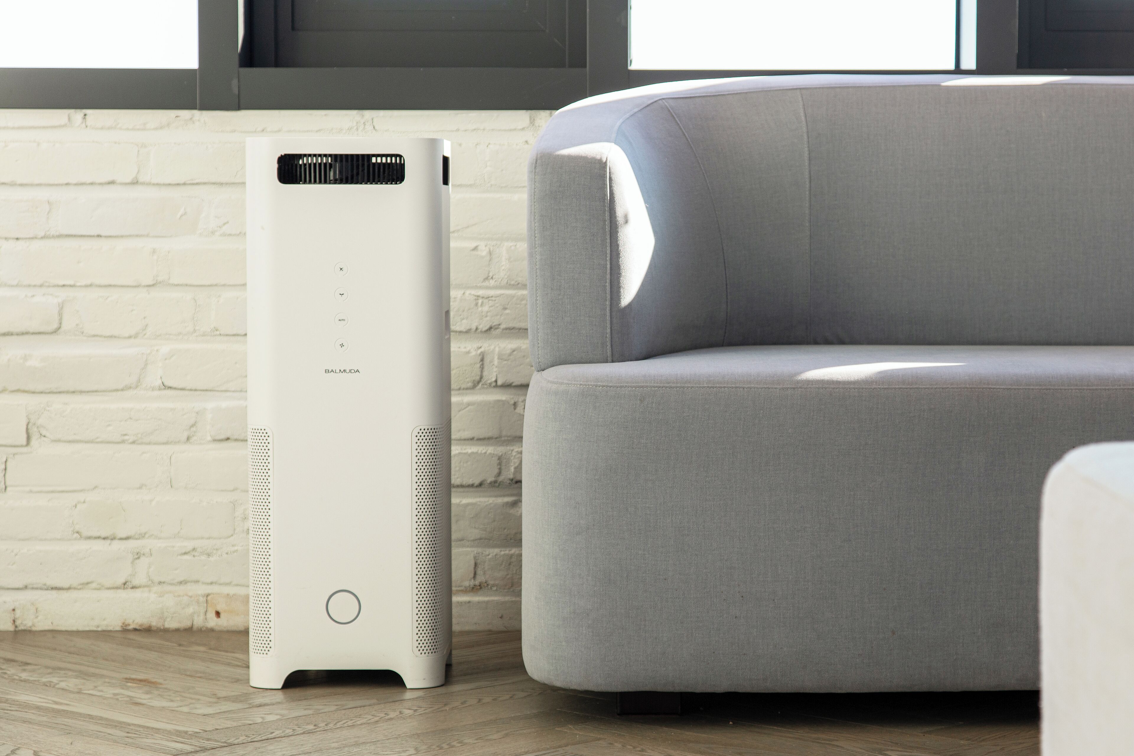 A white air purifier next to a grey sofa in a room with a white brick wall and a window.