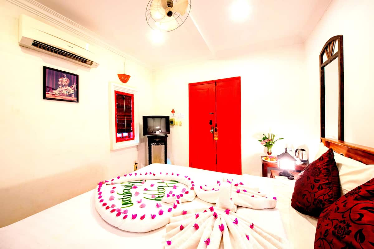 A hotel room with a bed, a red door, a small table with a lamp, and a wall-mounted air conditioner.