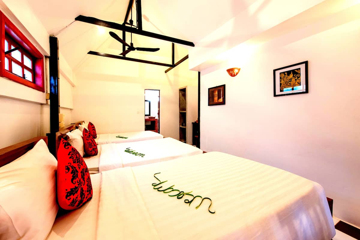 A hotel room with two beds, each with a white bedspread and a name embroidered in green. There is a red-framed window on the left and a framed picture on the wall.