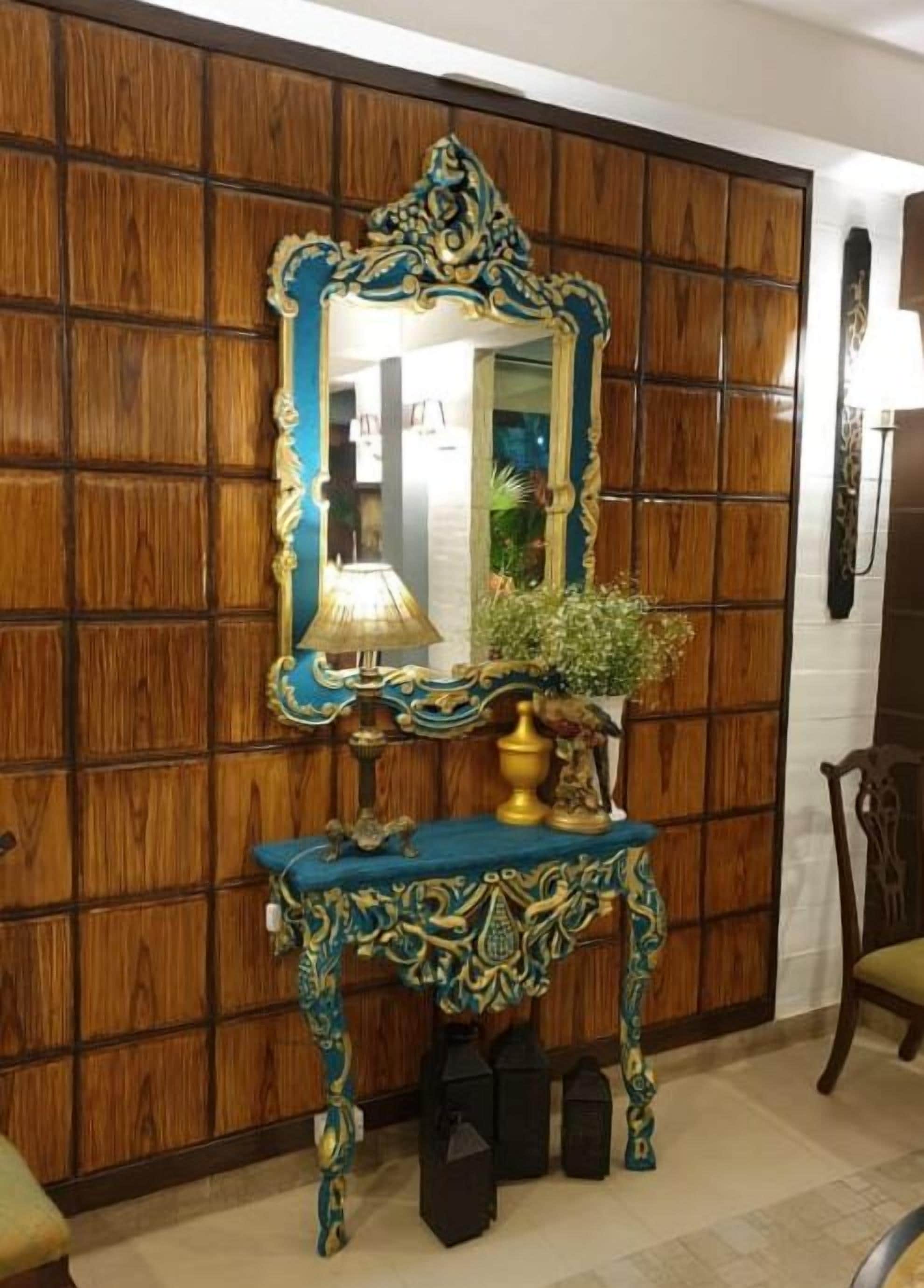 A room with a wooden panel wall, a decorative mirror, a blue console table with a lamp, and a potted plant.