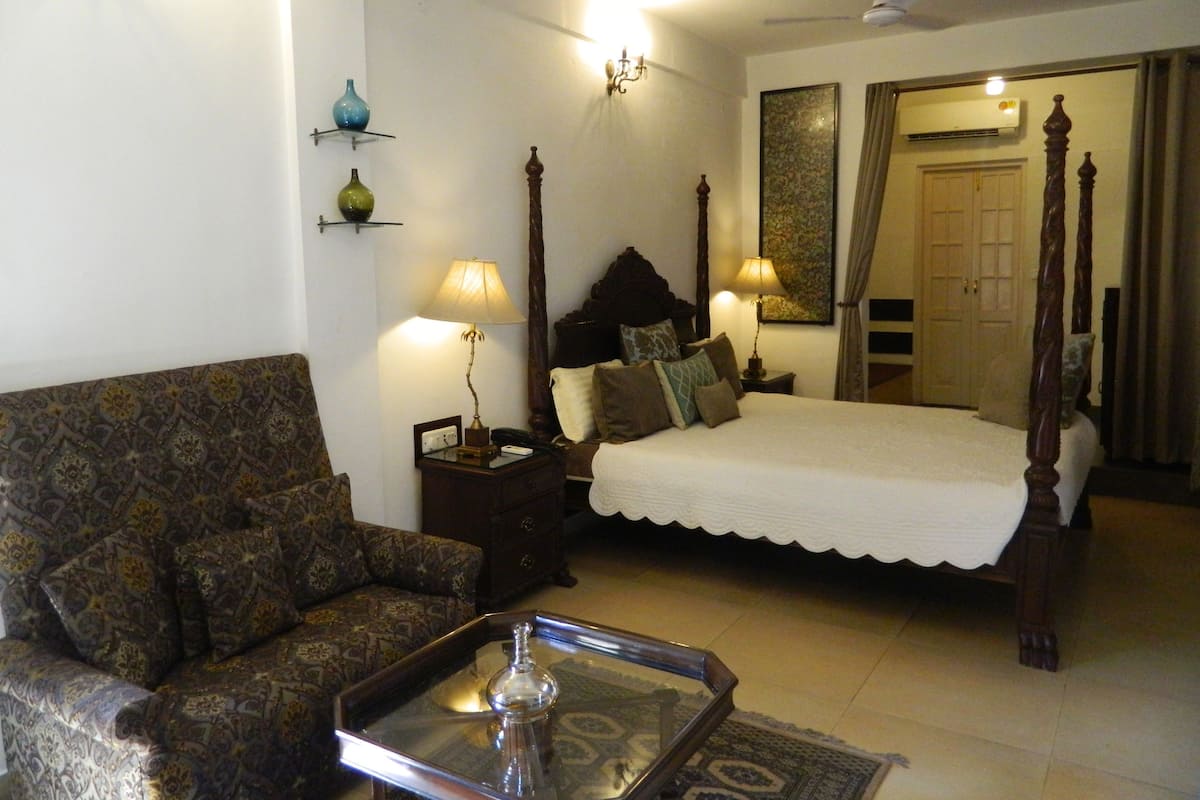 A bedroom with a four-poster bed, a nightstand, a lamp, a patterned sofa, and a decorative tray.