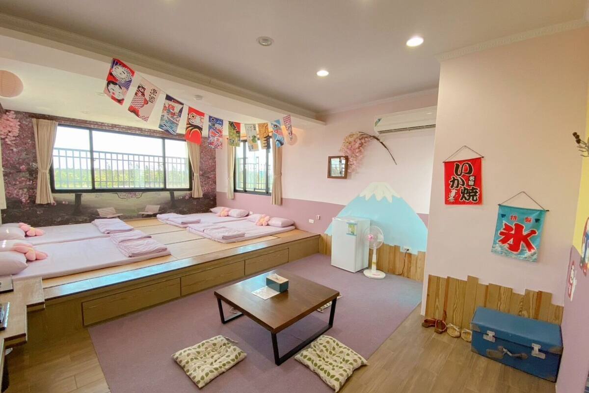 Una habitación con dos camas, una mesita, un ventilador y una ventana con cortinas.