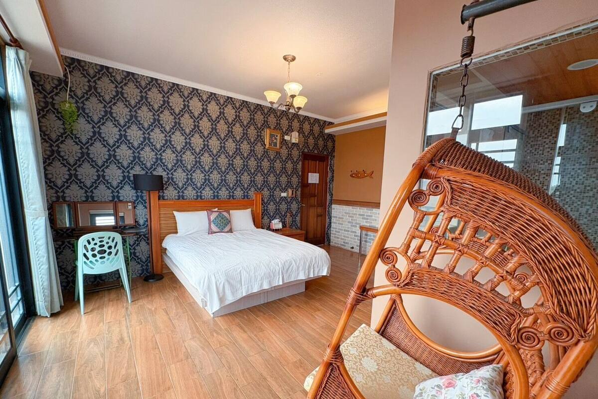 Un dormitorio con cama, una silla de mimbre, una mesita de noche, un espejo y un papel tapiz estampado.