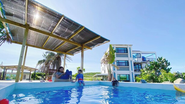 A swimming pool area with a shaded structure, palm trees, and a modern building in the background. - Big Fish House (Magong)