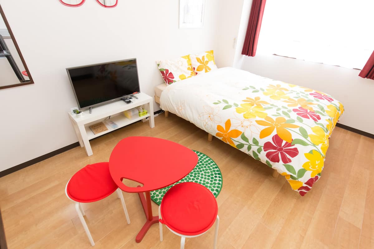 Un dormitorio con cama, televisor, una pequeña mesa con sillas y una ventana con cortinas.
