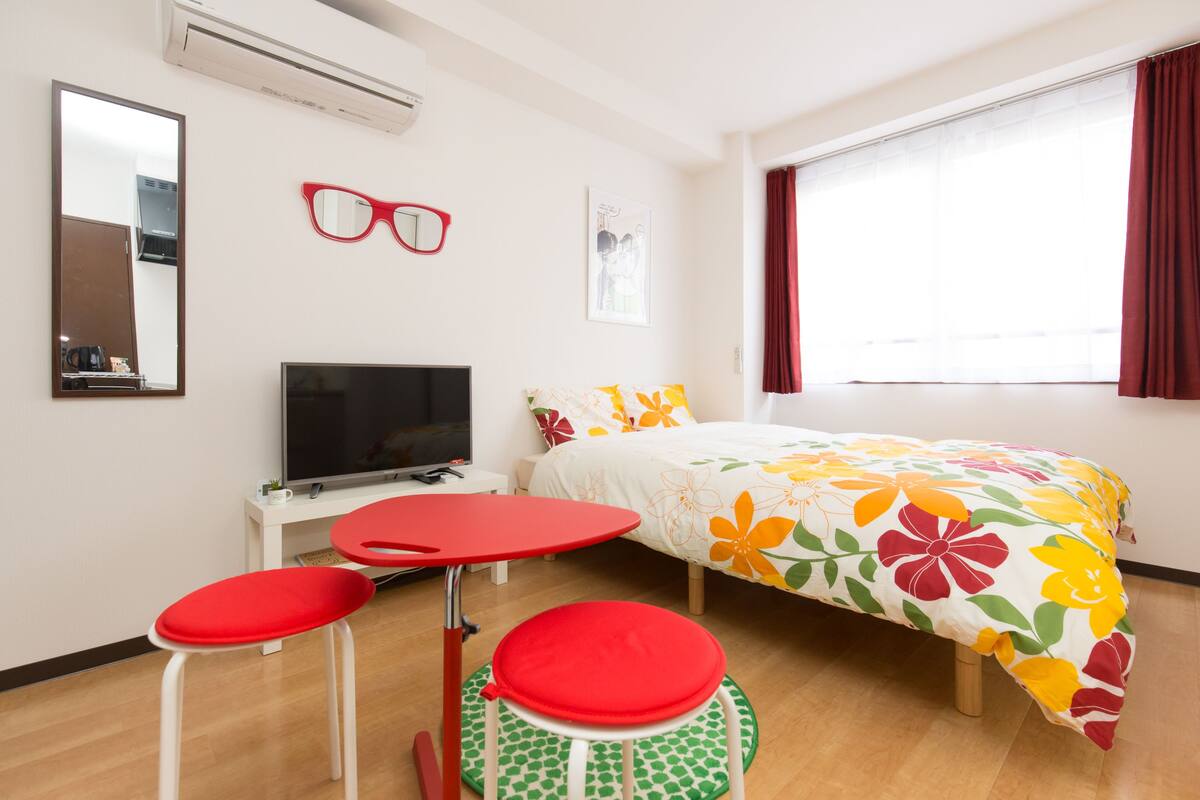 Una habitación de hotel con una cama, un televisor, un espejo y una mesa roja con sillas a juego.