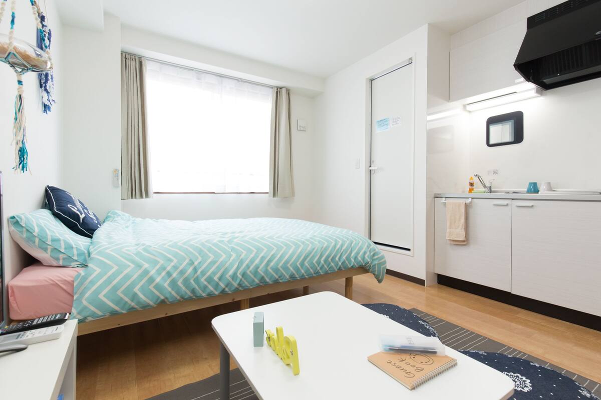 Habitación de hotel compacta con cama, escritorio pequeño, kitchenette y ventana con cortinas.