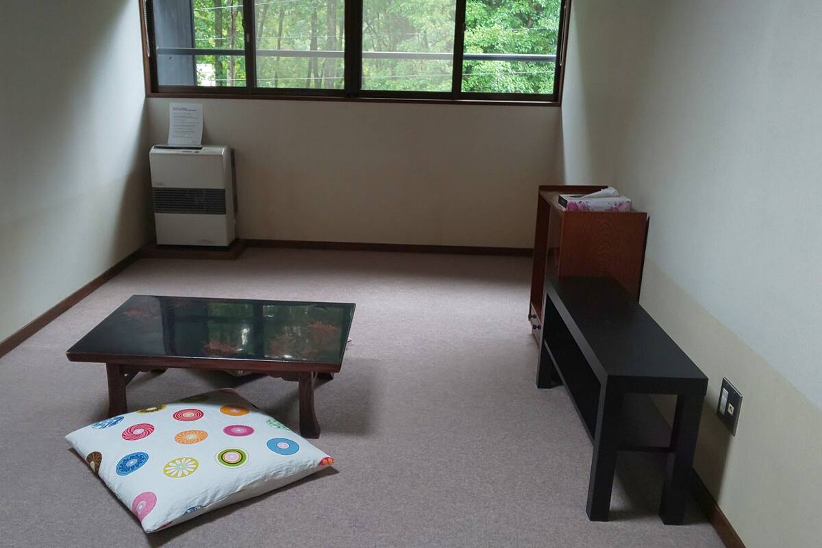 A small room with a glass-top table, a bench, a colorful cushion, and a window with a view of trees.