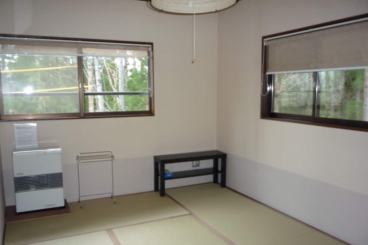 A room with tatami mat flooring, a standing heater, a small black table, and two windows with views of trees.