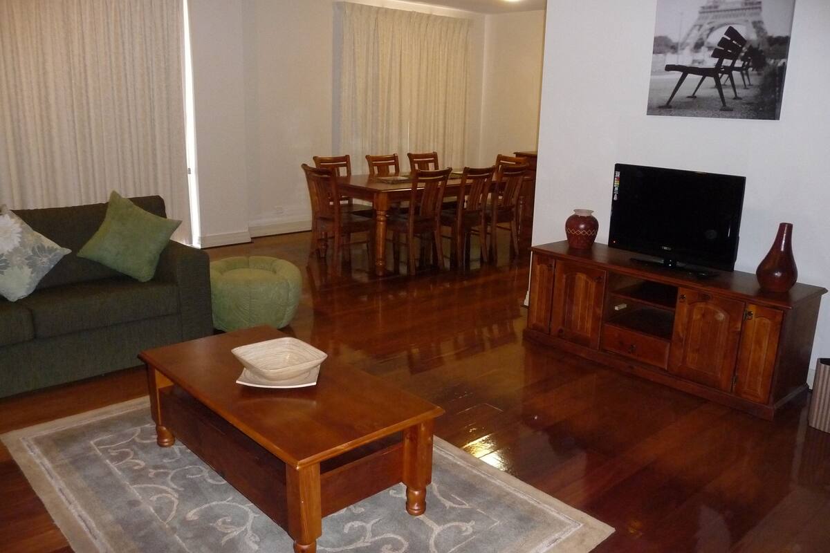 A living room with a green sofa, wooden coffee table, dining table with chairs, a TV on a wooden stand, and a framed picture on the wall.