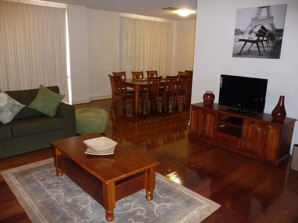 A living room with a green sofa, wooden coffee table, dining table with chairs, a TV on a wooden stand, and a framed picture on the wall. - City Escape 3BD in Adelaides East End 6 (Adelaide)