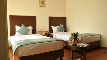 A hotel room with two beds, a small table with a fruit bowl and a drink, and a nightstand with toiletries.