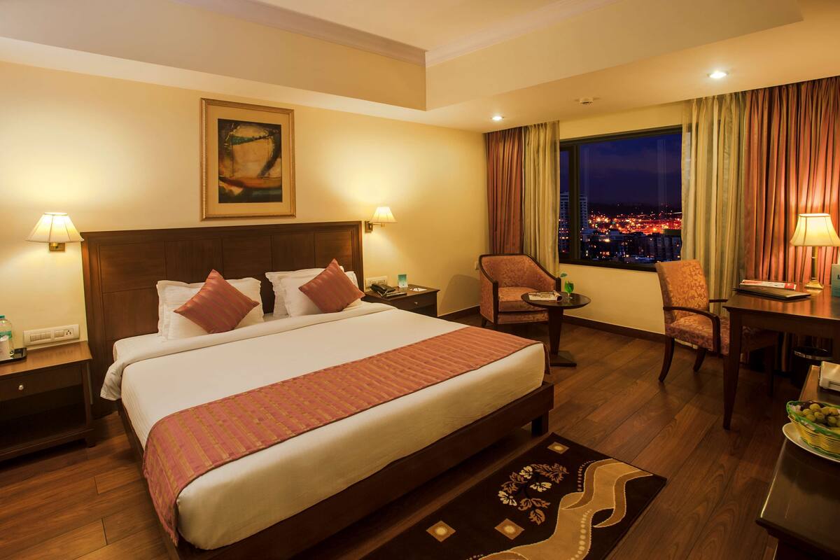 Habitación de hotel con una cama grande, un escritorio, una silla, una mesita de noche, una lámpara, un cuadro en la pared y vistas de la ciudad por la noche.
