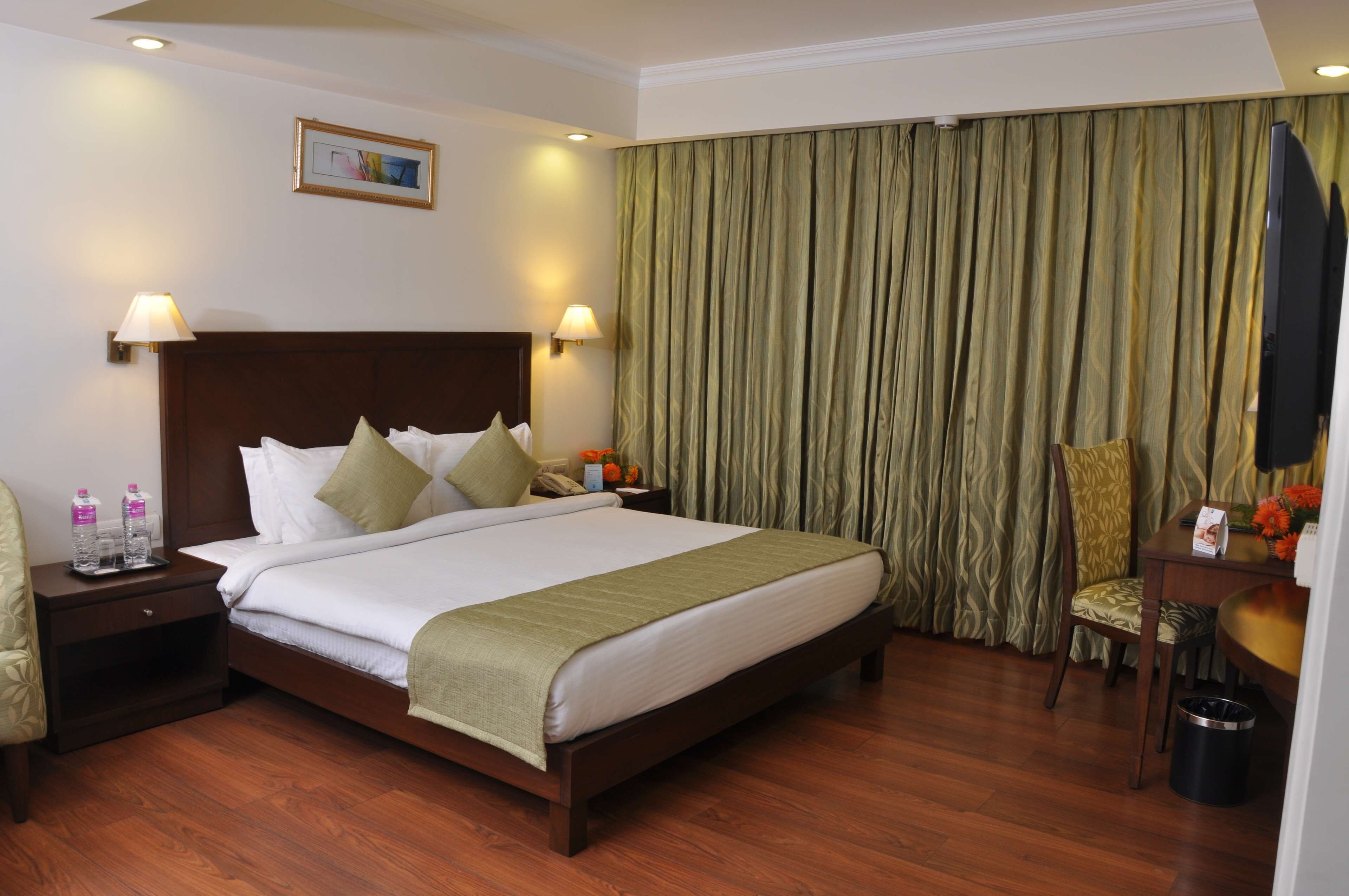 A hotel room with a large bed, two bedside lamps, a nightstand with a phone and water bottles, a desk with a chair, and a television.