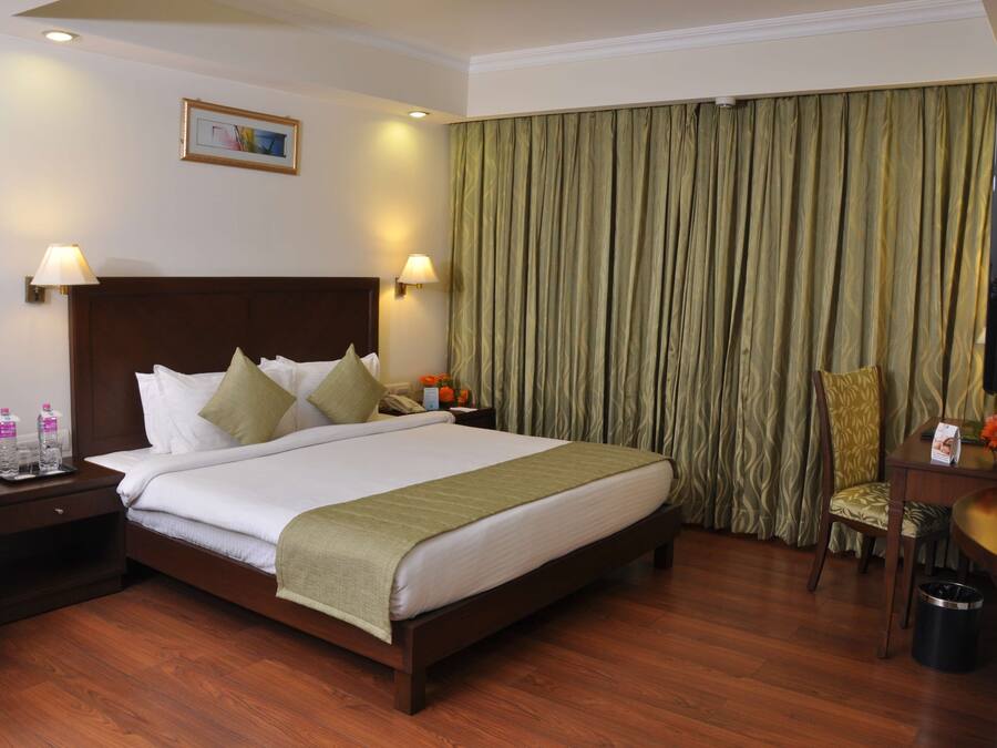 A hotel room with a large bed, two bedside lamps, a nightstand with a phone and water bottles, a desk with a chair, and a television.