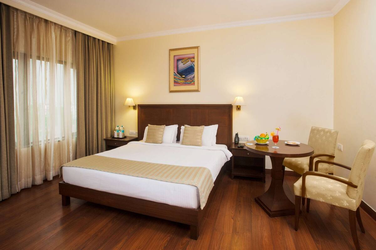 Habitación de hotel con una cama grande, un escritorio con silla, un cuadro en la pared y una ventana con cortinas.