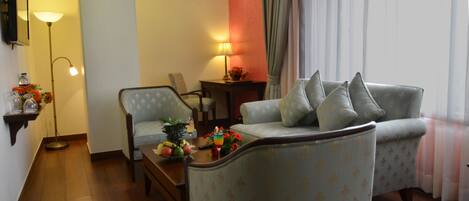 Ein Wohnzimmer mit einer Couch, Sesseln, einem Couchtisch mit Früchten und einem Getränk, einer Lampe und einem Fernseher an der Wand.