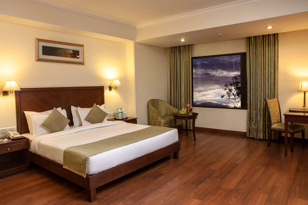 Habitación de hotel con una cama grande, suelo de madera, una mesita de noche con teléfono, una silla, una pequeña mesa con lámpara y una ventana con cortinas.