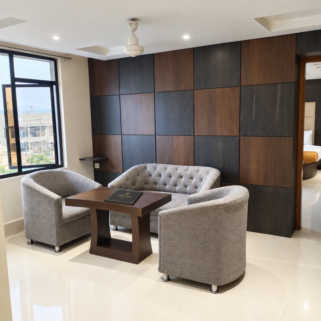 Una habitación de hotel moderna con un ventanal grande, un sofá capitonado, dos sillones, una mesita de madera y un panel decorativo en la pared.