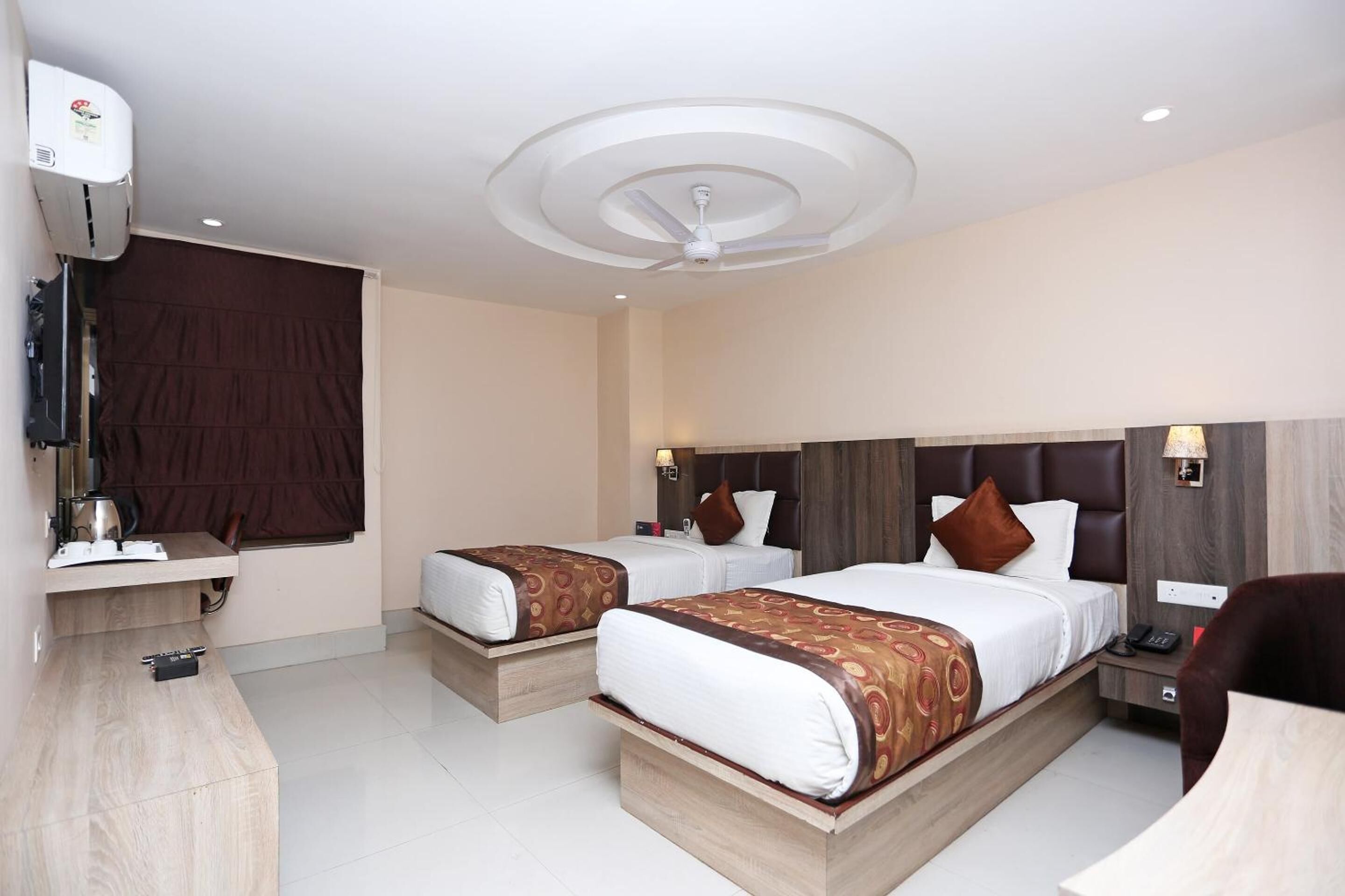 A hotel room with two beds, a desk, a chair, a ceiling fan, and a window with curtains.