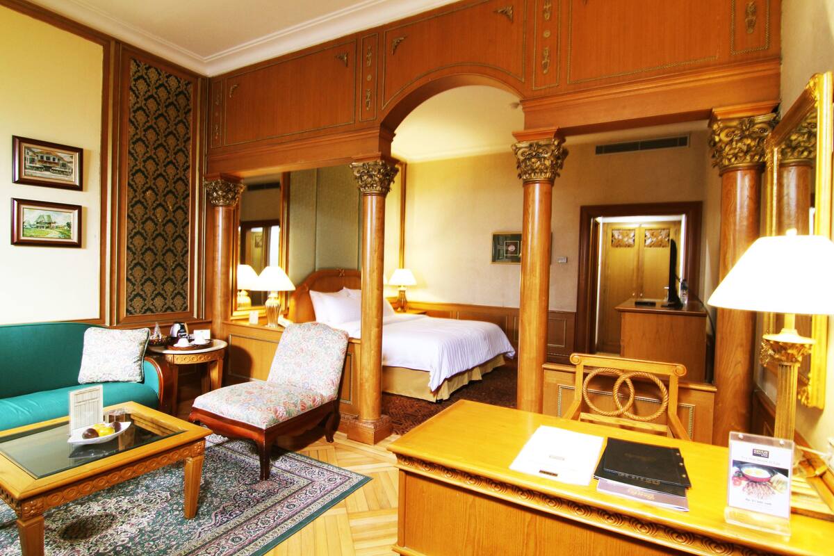 A hotel room with a bed, a sitting area with a sofa and armchair, a small table, and a desk with a computer.