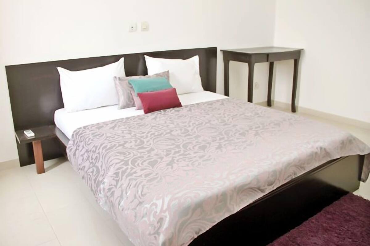 A neatly made bed with a patterned bedspread, a headboard, and multiple pillows. There is a bedside table with a lamp and a small nightstand with a phone.