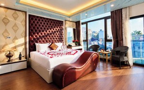 A hotel room with a large bed, a sitting area with a chair, a small table, and a view of the city through large windows.