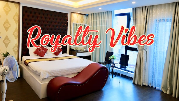 A modern hotel room with a large bed, a red upholstered headboard, a bedside table, a chair, a desk, and a window with curtains. - The Royal Hotel (Hanoi)