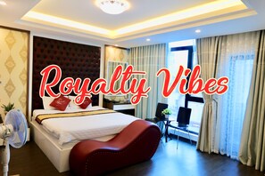 A modern hotel room with a large bed, a red upholstered headboard, a bedside table, a chair, a desk, and a window with curtains. - The Royal Hotel (Hanoi)