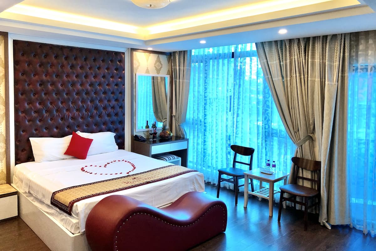 A hotel room with a large bed, a desk, a chair, and a view of the city through sheer curtains.