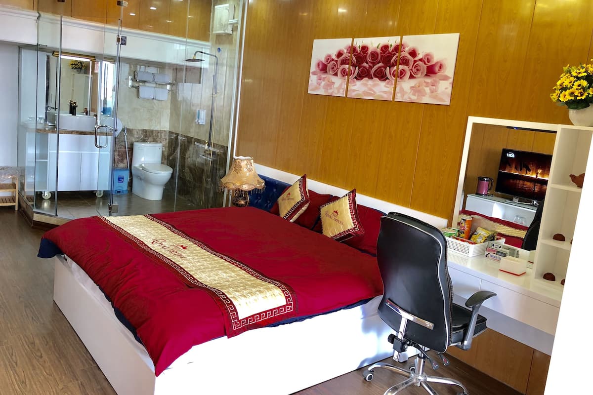 A hotel room with a large bed, a desk with a computer, a chair, a TV, and a bathroom with a glass shower enclosure.