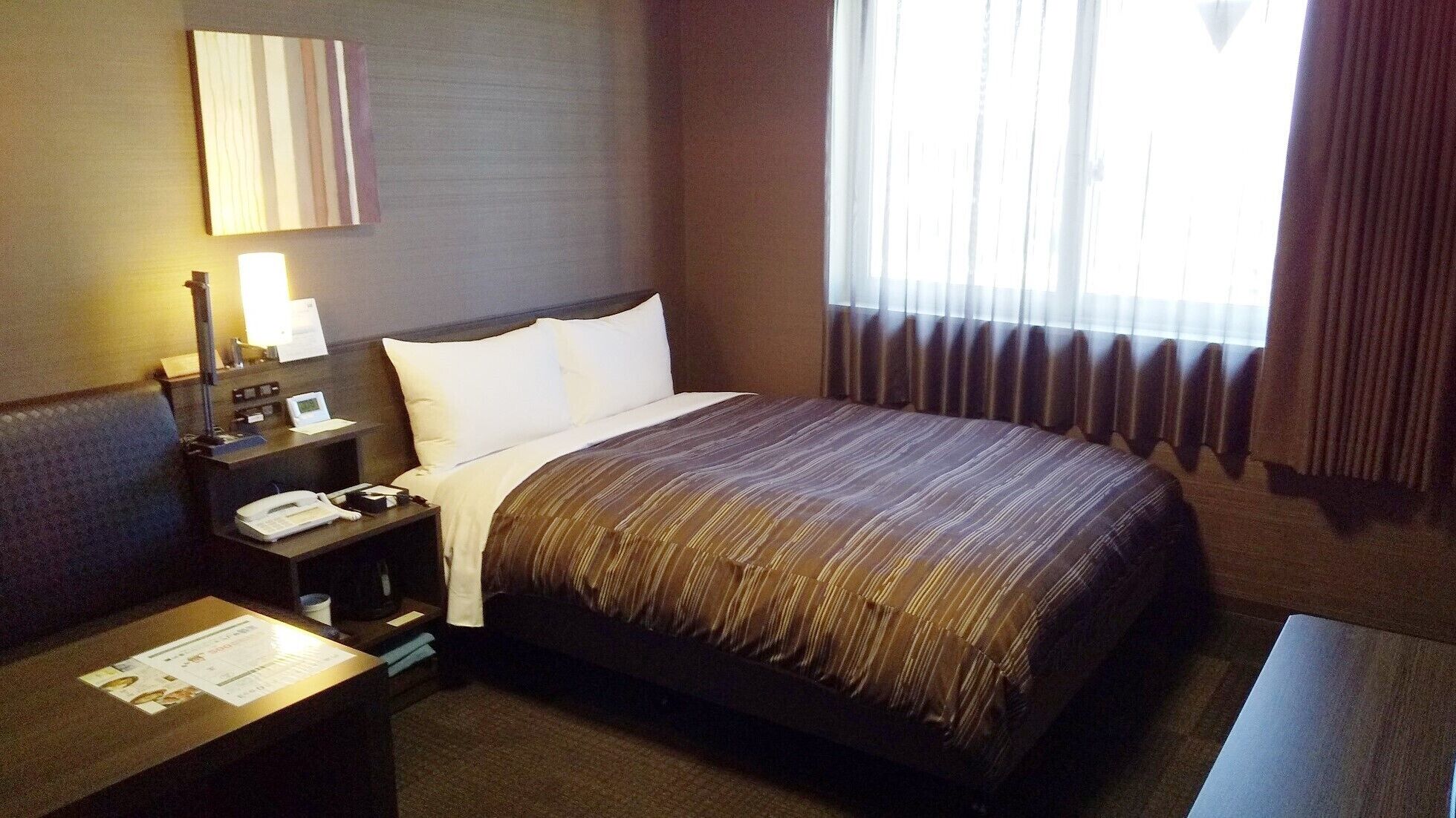 A hotel room with a bed, a nightstand with a phone and lamp, a window with curtains, and a wall-mounted mirror.