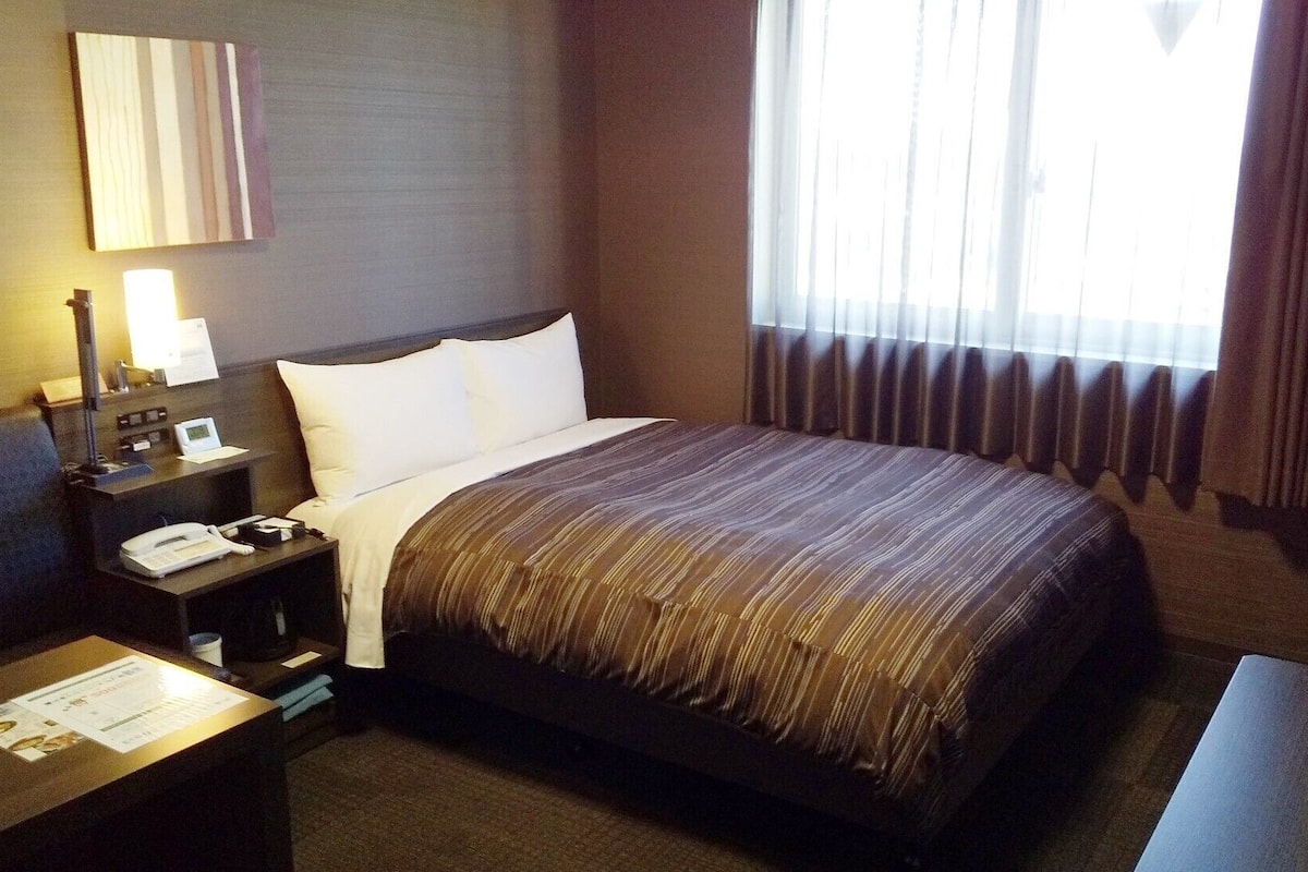 Ein Hotelzimmer mit einem Bett, einem Nachttisch mit Telefon und Lampe, einem Fenster mit Vorhängen und einem an der Wand befestigten Spiegel.