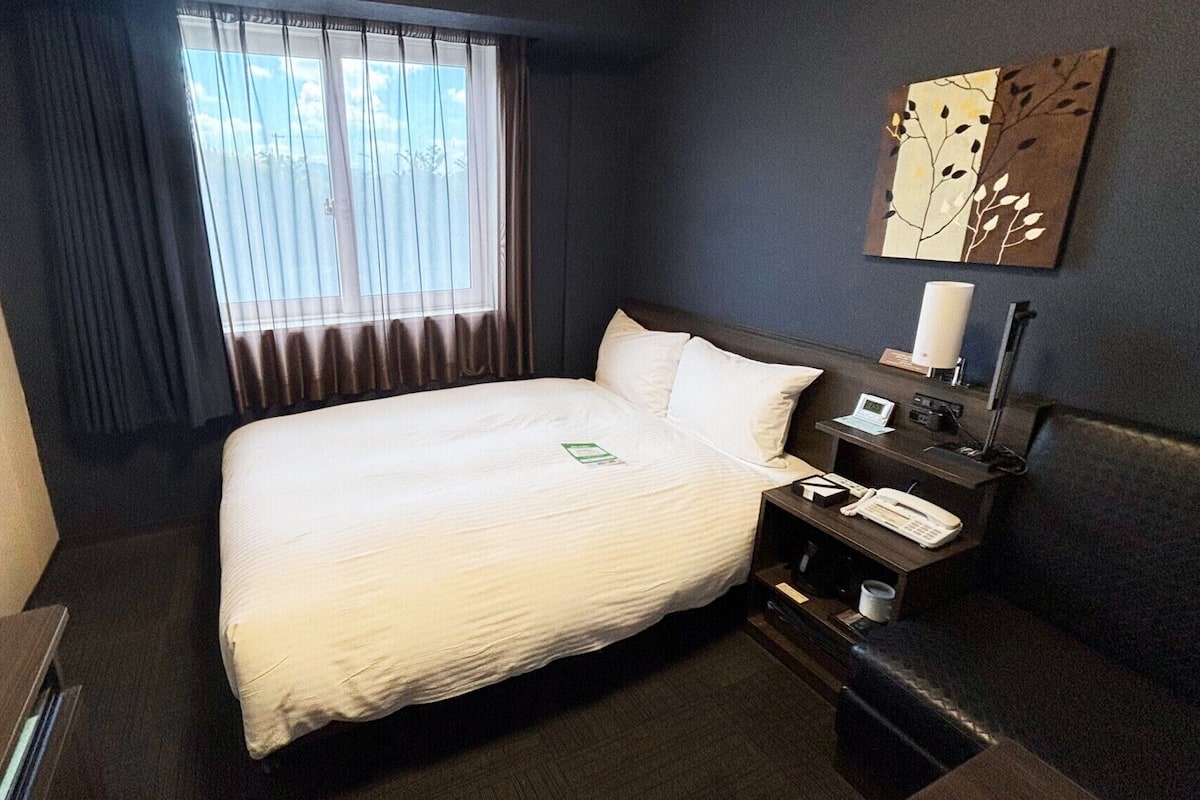 Ein Hotelzimmer mit einem großen Bett, einem Nachttisch mit Lampe, einem Telefon und einem Fenster mit Vorhängen.