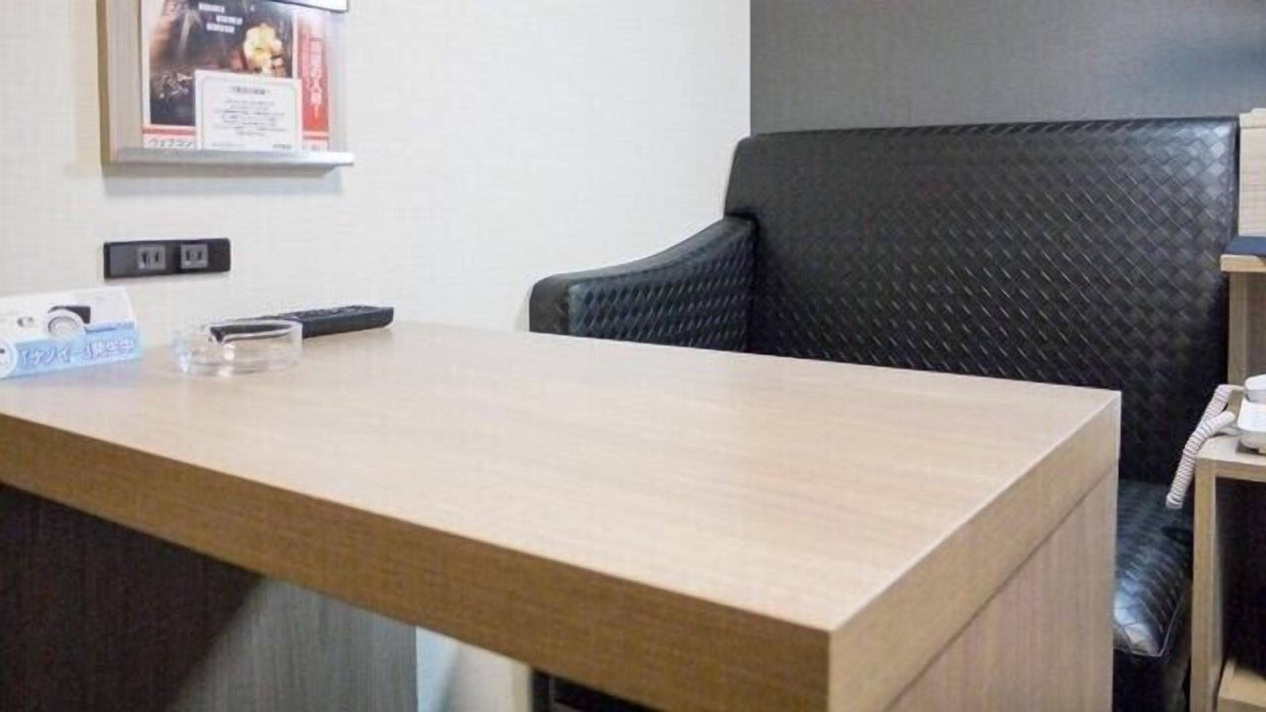 A hotel reception desk with a wooden surface, a black office chair, a framed certificate on the wall, and a telephone on a small table.