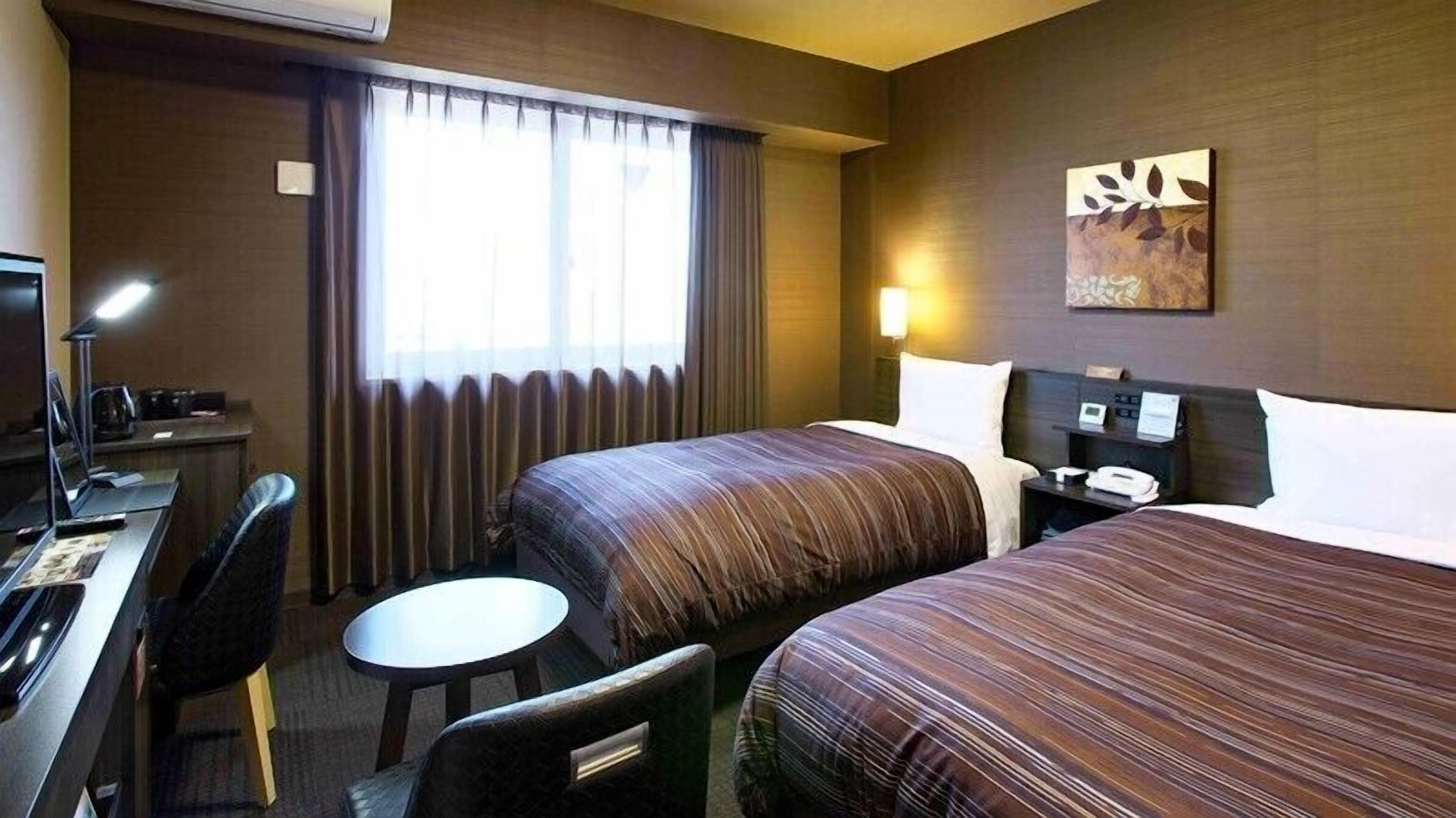 A hotel room with two beds, a desk with a chair, a small round table, a lamp, and a painting on the wall.