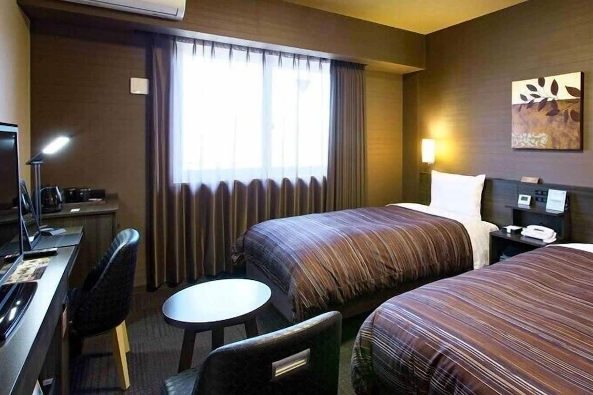 Ein Hotelzimmer mit zwei Betten, einem Schreibtisch mit Stuhl, einem kleinen runden Tisch, einer Lampe und einem Bild an der Wand.