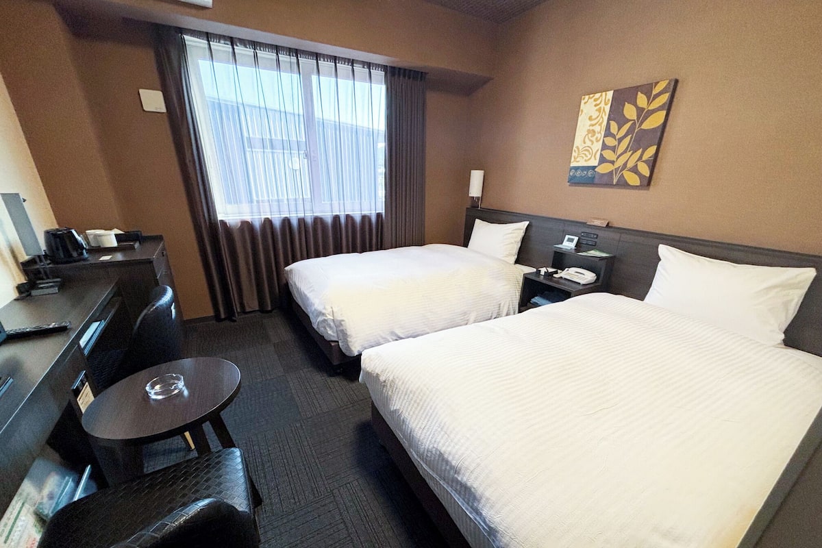 Ein Hotelzimmer mit zwei Betten, einem Schreibtisch, einem Stuhl, einem kleinen Tisch, einer Lampe und einem Fenster mit Vorhängen.
