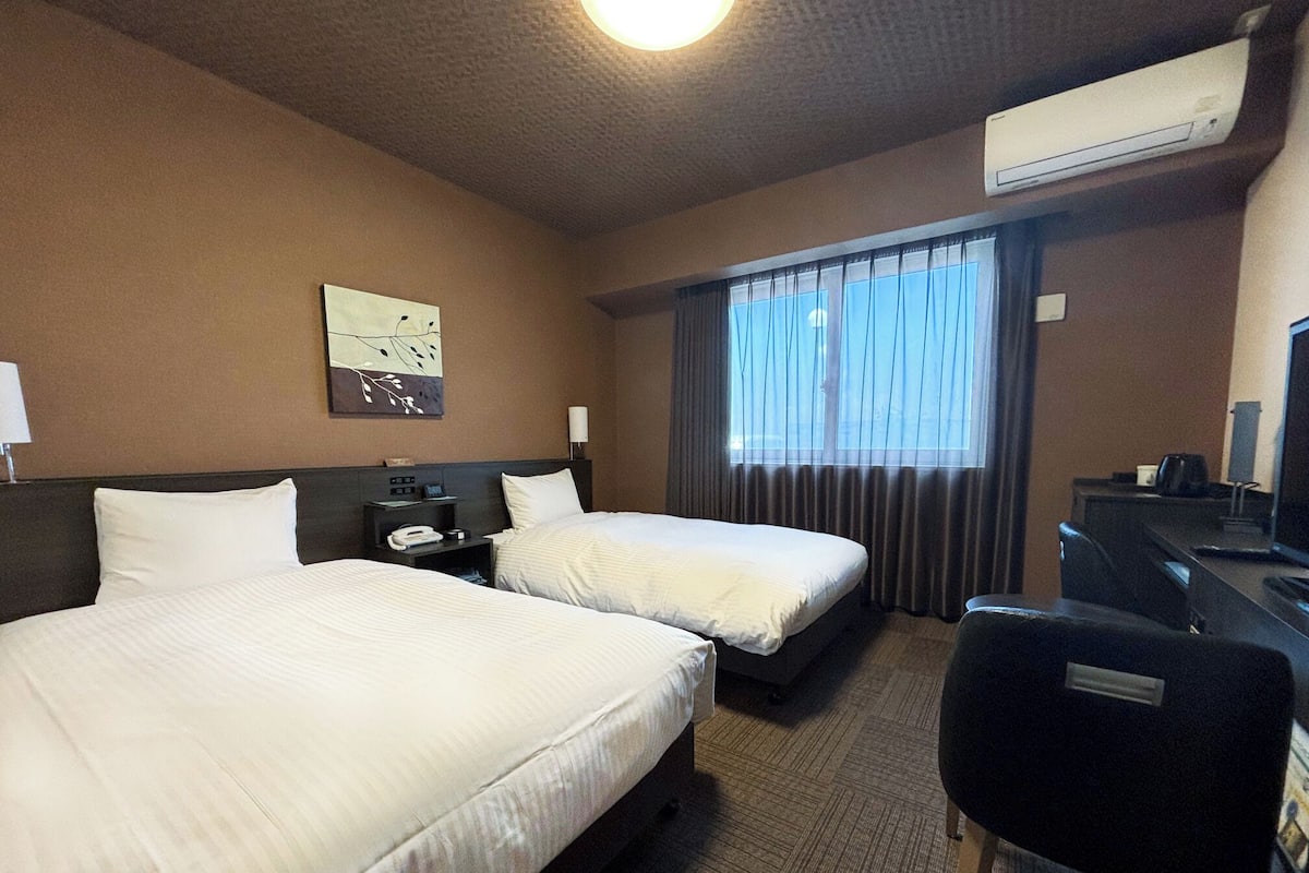 Ein Hotelzimmer mit zwei Betten, einem Schreibtisch, einem Sessel, einer Klimaanlage und einem Fenster mit Vorhängen.