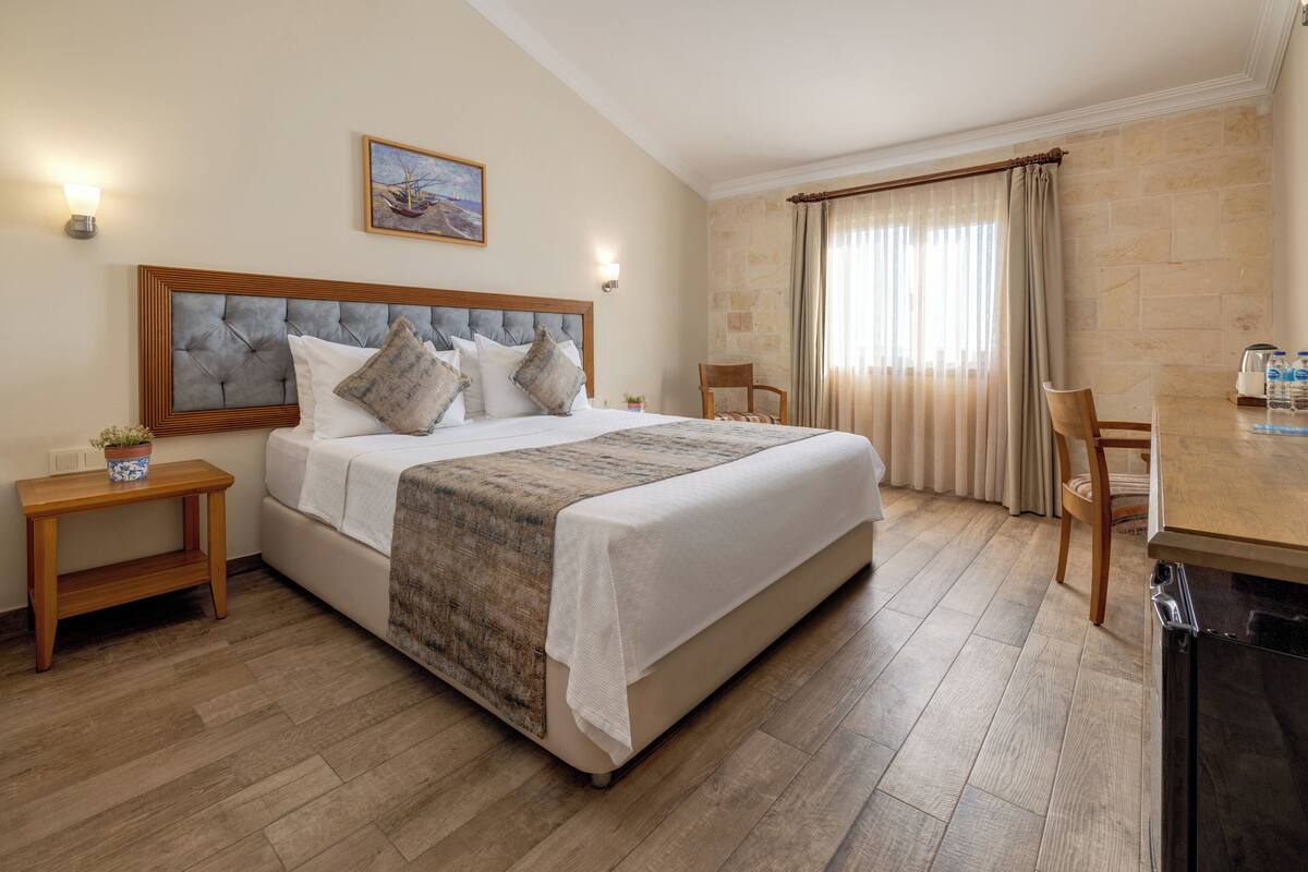 Quarto de hotel com uma cama grande, uma mesinha de cabeceira de madeira, uma pequena mesa, uma cadeira e vista para o mar pela janela.