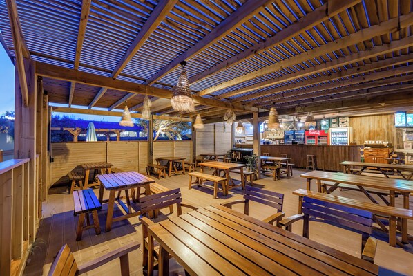 Una terraza exterior de madera con mesas de picnic y bancos, una máquina expendedora de Coca-Cola y una zona de bar con armarios de madera.