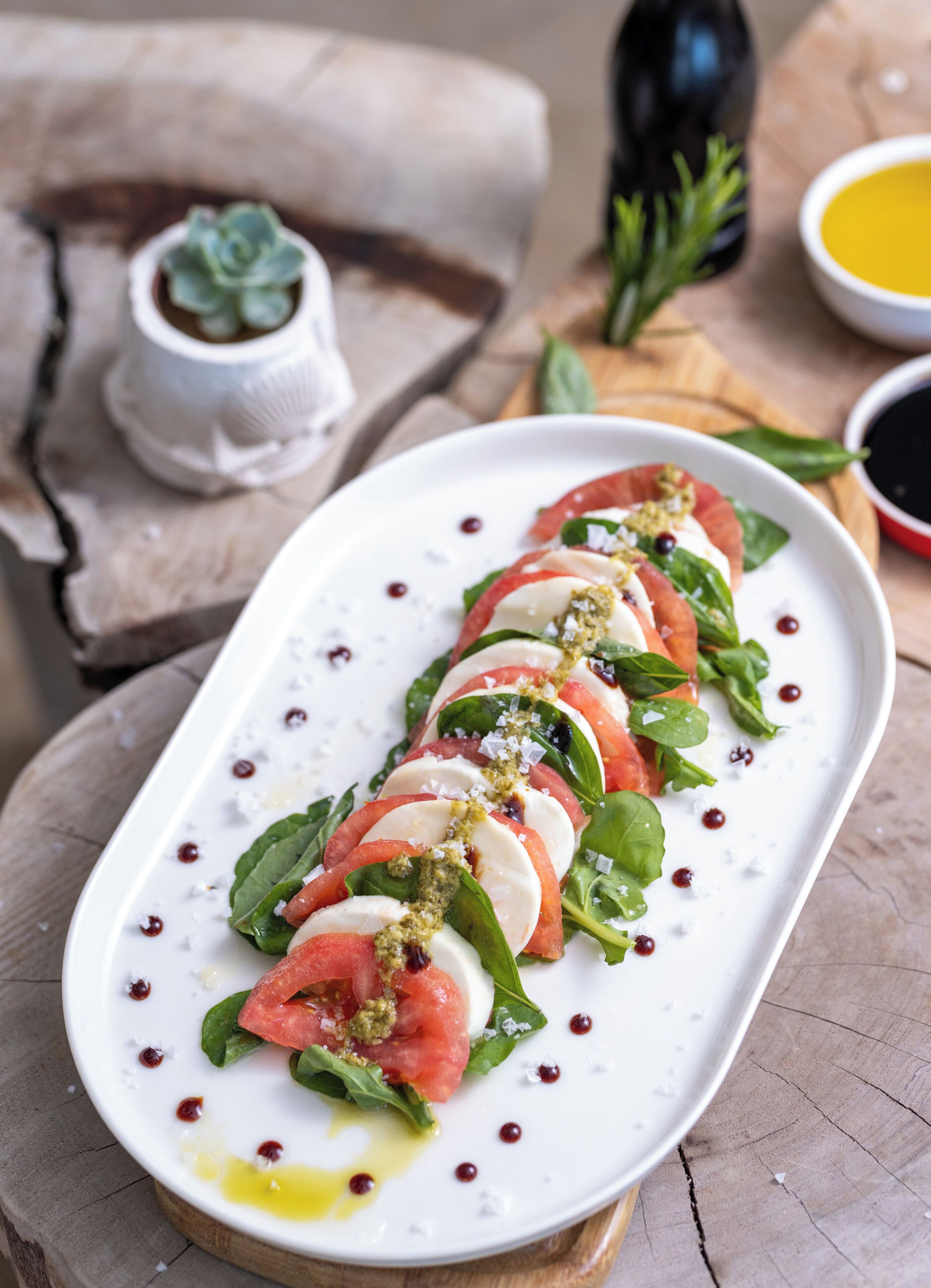 A white rectangular plate with a tomato, mozzarella, and basil salad, garnished with olive oil and salt, on a wooden table.